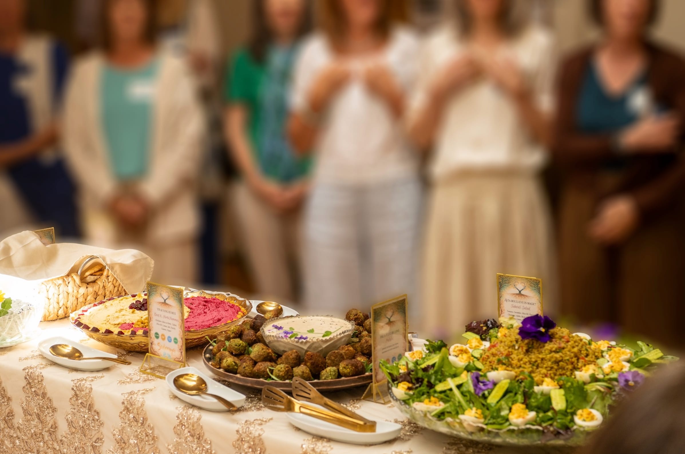 A beautifully arranged catering spread of hummus, falafel and flower-garnished salads on a sequined table, with a circle of guests in white gathered in ceremony softly blurred behind