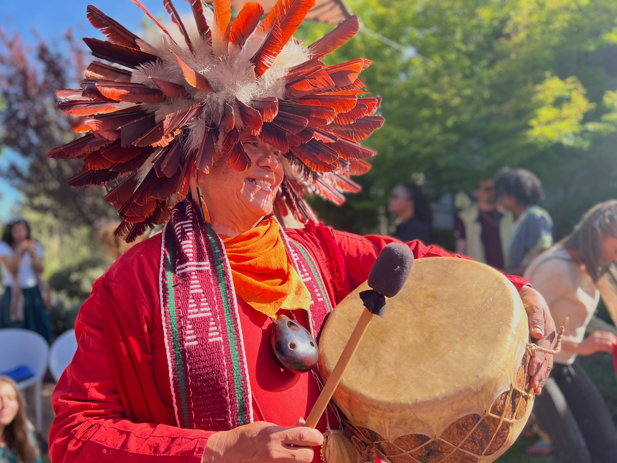 An elder in ceremonial red regalia and a feathered headdress playing a hand drum at a gathering