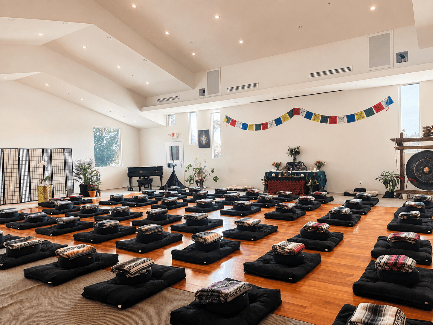 Meditation cushions in careful rows, piano, altar and gong waiting beneath prayer flags, set for an evening of sound healing and breathwork