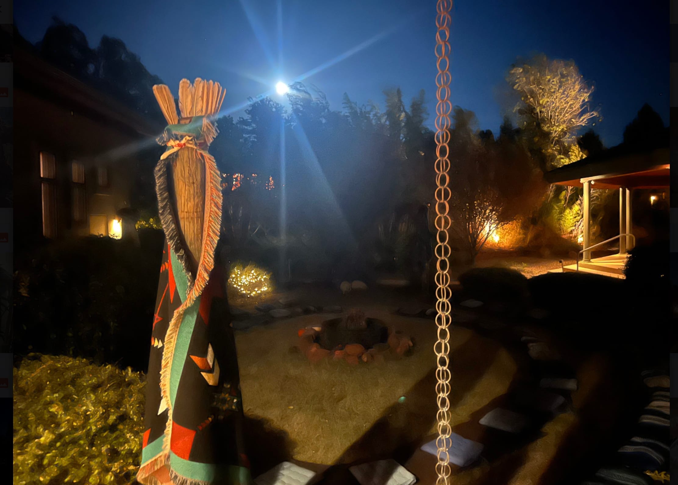 A feathered ceremonial staff catches the moonlight beside the tipi at dusk, the fire circle glowing beyond