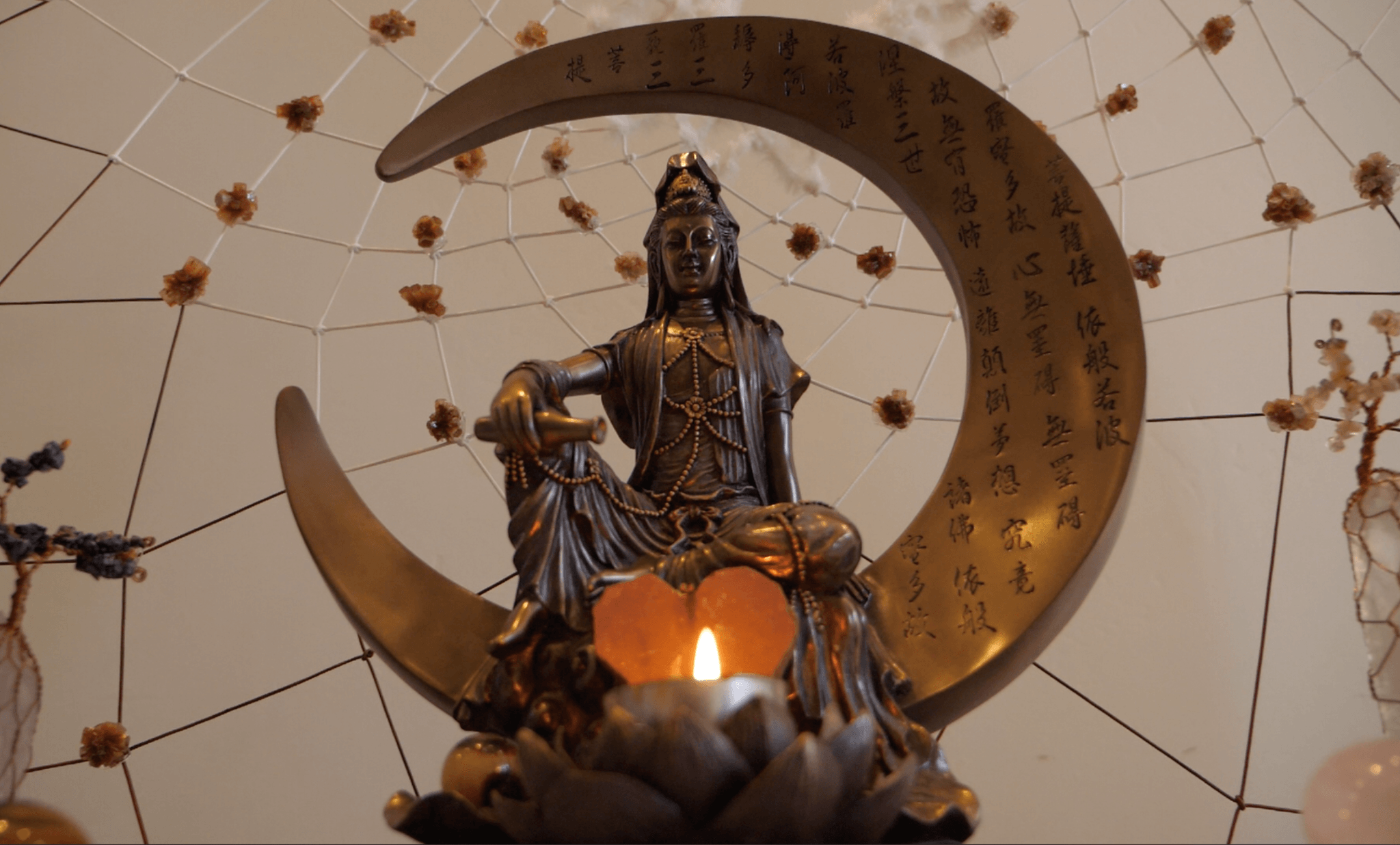 A bronze Guan Yin rests on a crescent moon inscribed with sutra, a single flame burning below